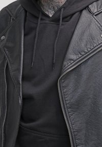Blouson motard en cuir noir avec zips argentés porté sur un sweat-shirt noir à capuche avec cordon de serrage et texture douce, mettant en avant un style superposé.