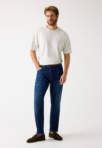 Lysegrå t-shirt med korte ærmer og logo, kombineret med mørkeblå lige ben bukser. Brune mokkasinstil sko fuldender outfittet.