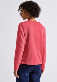 Koraalkleurige sweatshirt met lange mouwen, ronde halslijn en een textuurstof, gecombineerd met donkerblauwe jeans. Zichtbare stiksel en een casual pasvorm.