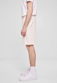 Homme portant un t-shirt blanc, un short beige clair jusqu'aux genoux, des chaussettes blanches et des baskets Nike blanches se tenant de côté contre un fond uni.