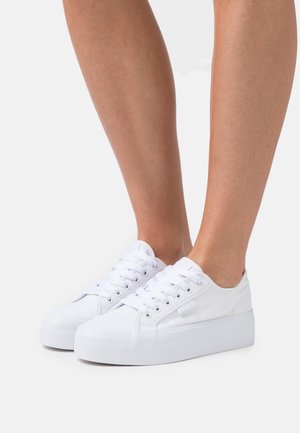 Personne portant des baskets blanches basses à plateforme avec des lacets, debout devant un fond blanc uni.