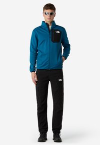 Giacca in pile verde acqua con zip e tasca sul petto nera, abbinata a pantaloni cargo neri. Stivali da trekking neri e occhiali da sole a specchio indossati.