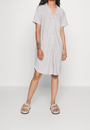 Frau, die ein knielanges, kurzärmeliges, vertikal grau-weiß gestreiftes Hemdkleid mit Brusttasche und beigen Sandalen trägt.