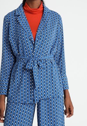 Femme portant une combinaison bleue à motifs ceinturée avec des manches longues, sur un haut à col roulé orange.