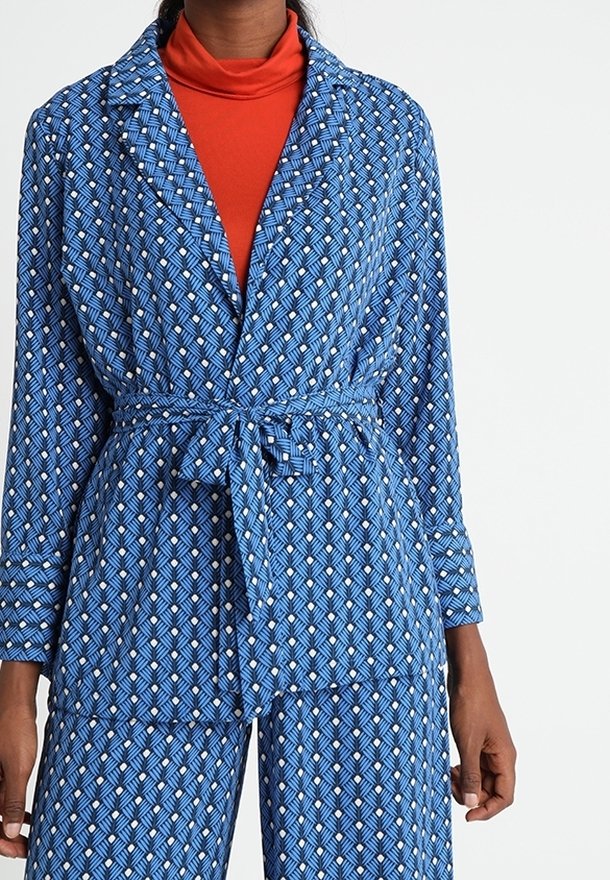Femme portant une combinaison bleue à motifs ceinturée avec des manches longues, sur un haut à col roulé orange.