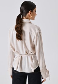 Blusa beige chiaro con maniche lunghe, spalle leggermente arricciate, vita legata e polsini con bottoni. Tessuto liscio, design del colletto classico.