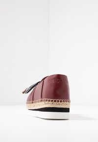 Espadrille en cuir rouge avec une semelle en jute tissée, dotée d'un lacet à motif noir et blanc et d'une marque en relief au niveau du talon.