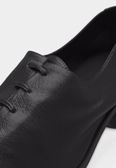 Μαύρο δερμάτινο παπούτσι με τακούνι, τρεις θηλιές για κορδόνια και λεία υφή, εμφανιζόμενο σε κοντινή λήψη από πλάγια και πάνω γωνίες.