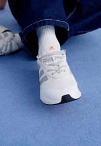 Bílé sportovní boty se šedými akcenty, šněrovací design, texturovaný svršek a černá podrážka. Nohou s bílými ponožkami; umístěno na modrém pozadí.