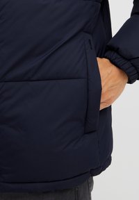 Marineblå puffjakke med en glatt, blank tekstur og elastiske mansjetter, med en sidelomme og en litt oversized passform.