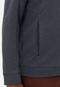 Giacca struttura in tessuto grigio scuro con tasca laterale e lacci. Presenta un orlo e polsini a coste che aggiungono struttura al design.
