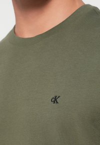 Nærbilde av en person som har på en olivengrønn genser med rund hals, med en liten svart "cK"-logo brodert nær brystet.