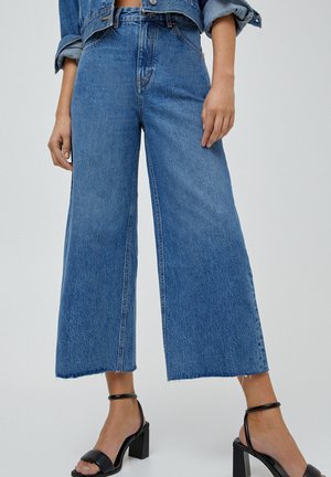 Donna che indossa jeans blu a vita alta e gamba larga con orli sfrangiati e sandali neri con cinturino alla caviglia e tacco largo, in piedi davanti a uno sfondo semplice.