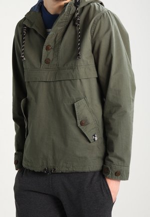 Mand iført en olivengrøn hættetrøje med knapdetaljer og en forlommer, kombineret med mørkegrå bukser.