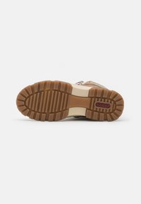 Baskets beiges avec surface texturée, semelle en caoutchouc présentant de profondes rainures et accents beige contrastants. Logo de la marque visible sur la semelle.