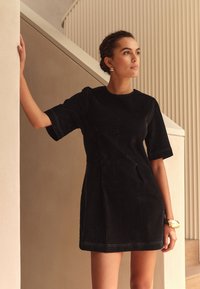 Crna haljina kratkih rukava s prilagođenim dizajnom i vidljivim šavovima. Glatka tekstura i dužina do koljena, uparen s velikim zlatnim narukvicama.