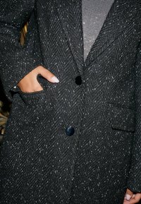 Sort hørfisket coat med glossy knapper, tekstureret stof og sidelomme. Synlig indre lag har et mesh-design.