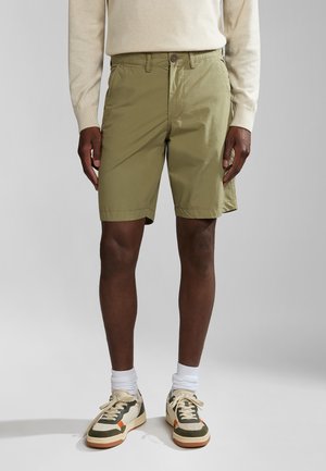 Homme portant un short vert olive, un haut manches longues beige, des chaussettes blanches et des baskets vertes, beiges et orange, debout sur un fond uni.