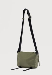 Τσαντάκι ώμου από ύφασμα σε ελιά πράσινο χρώμα με μαύρο ρυθμιζόμενο λουράκι, ορθογώνιο σχήμα, κλείσιμο με φερμουάρ και διπλά διακοσμητικά λουράκια στο κάτω μέρος.