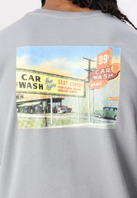 Grauer Pullover mit einem Vintage-Autowaschgrafik auf der Rückseite, die eine Beschilderung in Gelb und Rot mit einer klassischen Auto-Illustration zeigt.