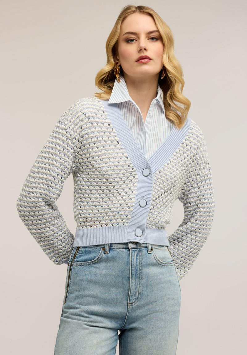 Mujer con cabello rubio ondulado que lleva grandes pendientes dorados, un cárdigan de punto azul y blanco con botones, una camisa a rayas azul clara y jeans de talle alto.