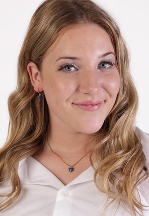Gouden ketting met een blauwe hartvormige hanger. Het model draagt ook bijpassende gouden ringoorbellen. Witte blouse met kraag en knoopdetail.