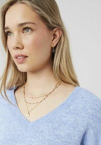 Pull en tricot bleu clair avec un col en V. Trois colliers en or superposés avec des accents noirs ; petites boucles d'oreilles circulaires avec un design de compas.