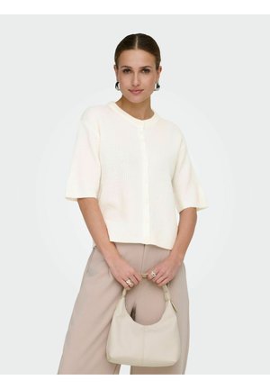 Vrouw draagt een wit, kort mouwens vest met knopen en beige broek, houdt een crèmekleurige handtas met beide handen voor zich vast.