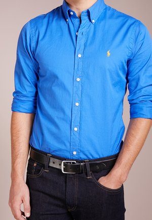 Camisa azul de botones en mezcla de algodón con mangas largas, presenta un cuello puntiagudo y un pequeño logo en el pecho, combinada con jeans oscuros y cinturón negro.