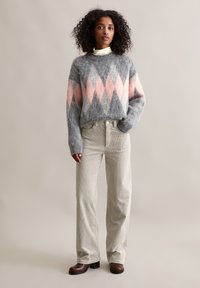 Grauer Strickpullover mit pink-grauem Rautenmuster, kombiniert mit beigen Cordhosen. Model trägt braune Stiefel mit Absatz.