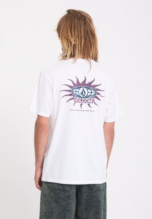Personne aux cheveux mi-longs portant un t-shirt blanc avec un soleil violet et le logo Volcom dans le dos, associé à un short foncé.