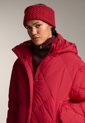 Rote Puffjacke mit rautenförmigem Steppmuster und Kapuze, begleitet von einem textured roten Haarband. Darunter befindet sich ein dunkelroter Rollkragenpullover.