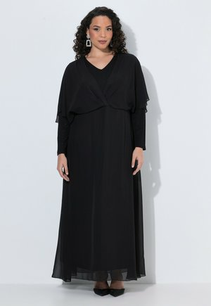 Femme aux cheveux bouclés portant une longue robe noire, un châle noir transparent, des boucles d'oreilles scintillantes et des chaussures noires pointues, debout devant un fond uni.
