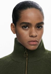 Maglione a collo alto in verde oliva a coste, con una zip dorata e dettagli con logo, caratterizzato da un tessuto testurizzato e un design aderente.