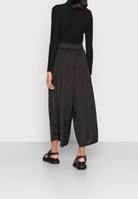 Sort ribbet langærmet top med plisserede sorte vide bukser. Bukserne har en høj talje og en glat tekstur. Sorte sandaler er synlige.