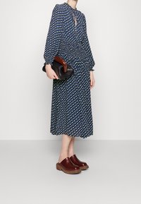 Robe midi bleu marine avec un motif de diamants blancs et jaunes, taille froncée et manches longues. Associée à une pochette noire et des sabots bordeaux.