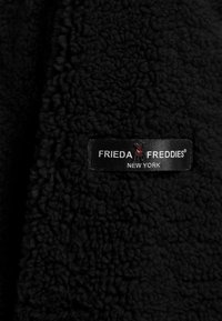 Zwarte fleece stof met een gestructureerd, pluche oppervlak. Bevat een zwart label met witte tekst en een logo, waarop staat "FRIEDA FREDDIES NEW YORK."