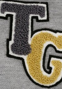 Szary materiał z teksturowanym haftem liter "T" w fioletowym i "G" w żółtym, obramowany czarnym i białym przeszyciem.