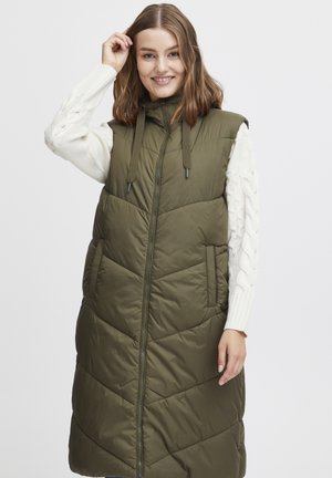 Gilet matelassé vert olive avec fermeture éclair, doté d'une capuche, de poches sur les côtés et d'un design en quilting. Porté par-dessus un pull en laine torsadée crème.