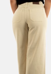 Jeans de pierna ancha beige con cintura alta, que cuentan con dos bolsillos traseros y costuras visibles. La tela tiene una textura suave y resistente.