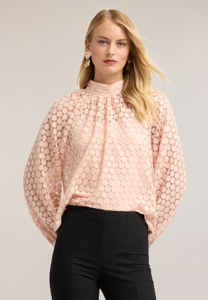 Donna con lunghi capelli biondi, che indossa grandi orecchini dorati, una camicetta di pizzo rosa pallido con maniche a sbuffo e pantaloni neri a vita alta, rivolta in avanti.