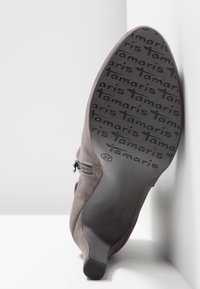 Botte à talon en daim avec fermeture éclair, montrant le motif de la semelle répétant "Tamaris" et la taille 37 marquée près de la voûte plantaire.