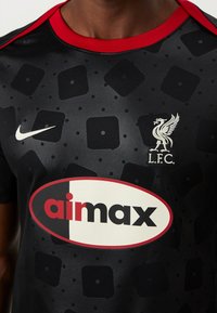 Musta lyhythihainen Nike-paita, jossa on punainen kaulus ja suuri valkoinen "aimax"-logo sekä teksturoidut neliökuviot koko paidassa. Mukana Liverpool FC:n tunnus.