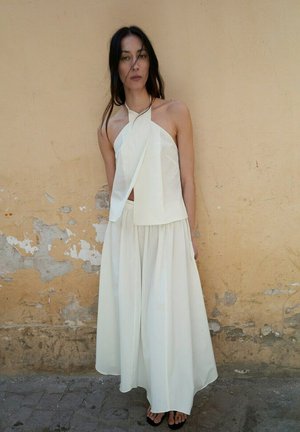 Femme aux longs cheveux foncés portant un haut blanc sans manches à col halter et une jupe blanche fluide devant un mur beige usé.