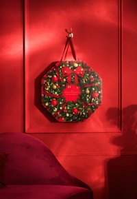 Achtseitiger Adventskalender mit festlichem Design, das rote und goldene Ornamente, Grünpflanzen und eine zentrale Schleife umfasst, an einer roten Wand montiert.