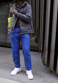 Giubbotto puffo grigio scuro, pantaloni della tuta blu con dettagli neri, sneaker bianche e una borsa a spalla verde. In piedi su una superficie tessurizzata.