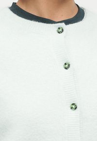 Κοντινό πλάνο σε λευκή πλεκτή ζακέτα με τρία πράσινα και μαύρα μαρμάρινα κουμπιά πάνω από σκούρο ριμπ κασκόλ.