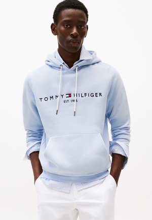 Junger Mann trägt einen hellblauen Tommy Hilfiger Hoodie über einem gestreiften Hemd und weißen Hosen, steht mit den Händen in den Taschen vor einem einfarbigen Hintergrund.