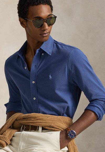 Μπλε πουκάμισο μακρυμάνικο με ανοιχτό μπλε λεπτομέρειες, λευκά κουμπιά και μικρό λογότυπο. Συνδυάζεται με μπεζ πουλόβερ δεμένο γύρω από τη μέση και ρολόι.