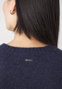 Femme aux cheveux raides et foncés portant un pull bleu marine avec un petit logo métallique "BOSS" au niveau de la nuque,  à l'arrière, et une boucle d'oreille argentée en forme d'anneau.